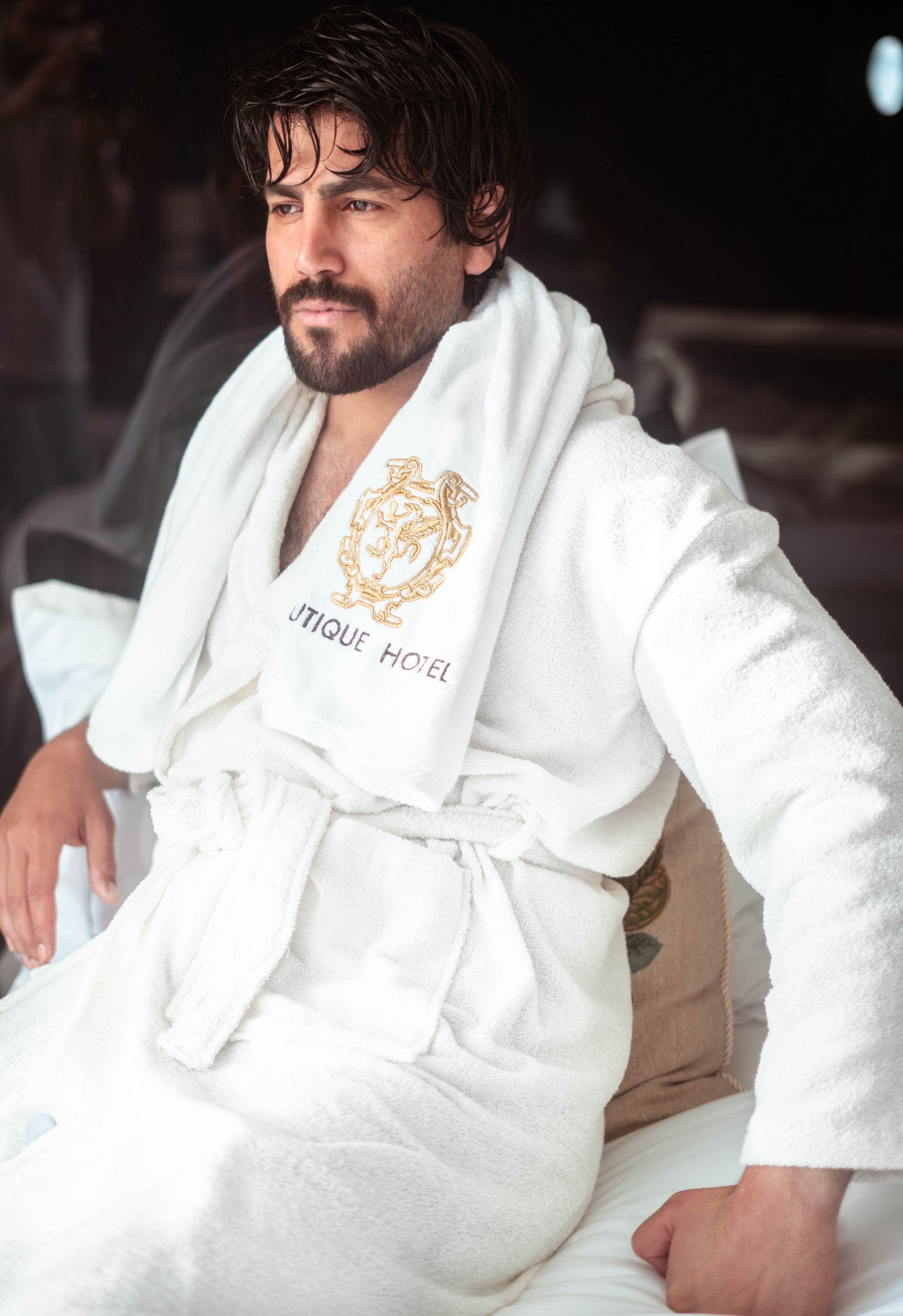 Man in a plush white hooded bathrobe sitting on a bed in soft natural light