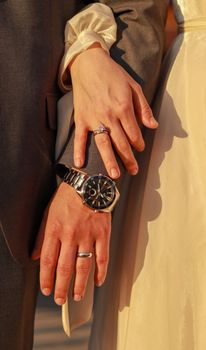 Close-up of a couple's hands with rings and a watch, warmly lit by the sun.