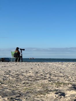 Solo photographer sits on beach observing ocean with telephoto lens on a sunny day.