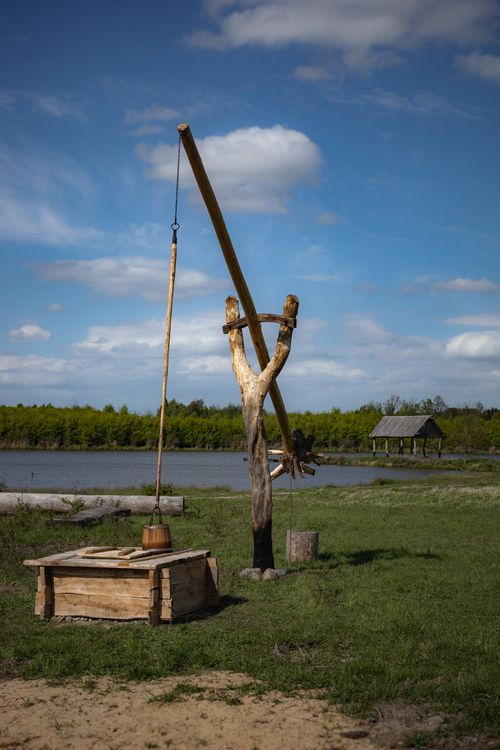 Free A traditional wooden well stands beside a peaceful lake under a clear blue sky. Stock Photo