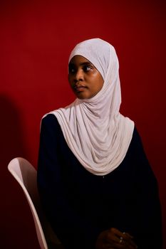 Elegant portrait of a woman in a white hijab sitting against a bold red background.