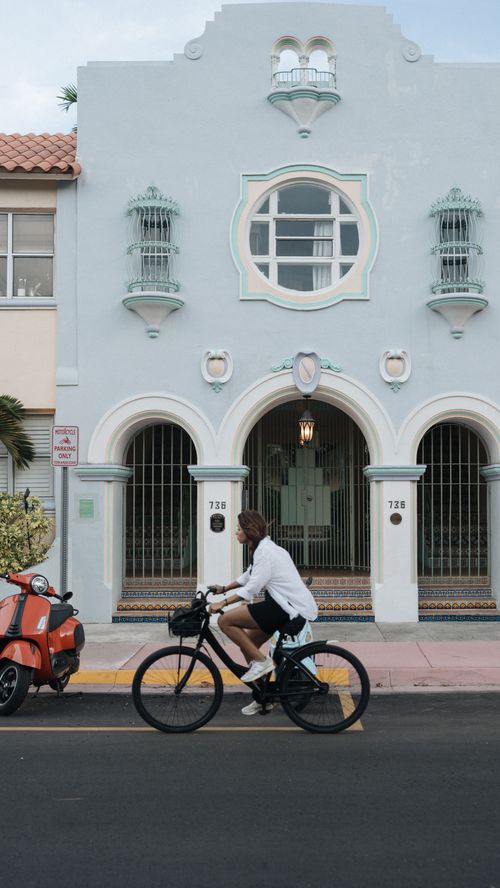 Free Man cycling past iconic Art Deco architecture on a sunny day, vibrant urban scene. Stock Photo