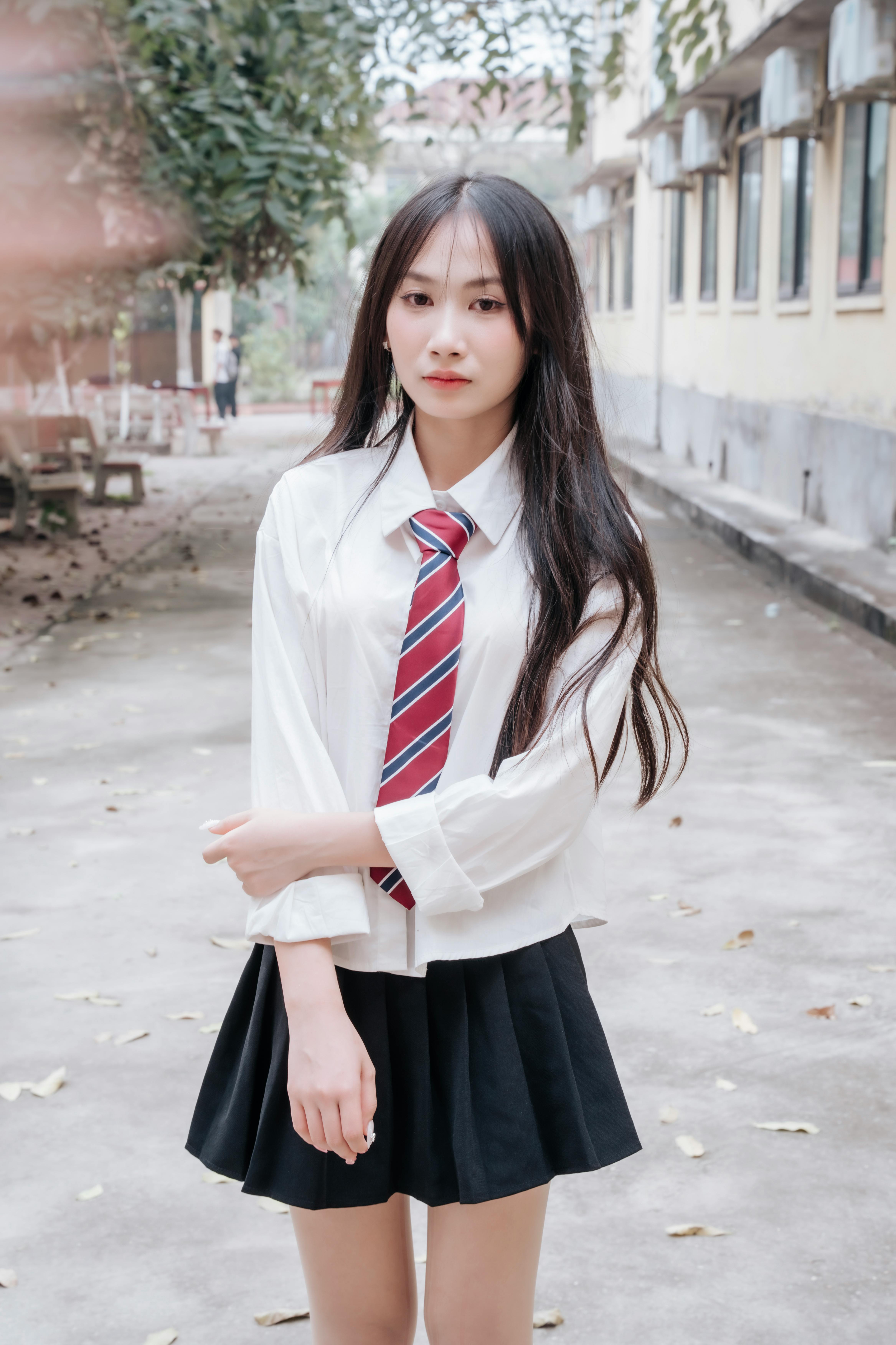 A young woman poses in a school uniform with a tie outdoors on a tree-lined path.