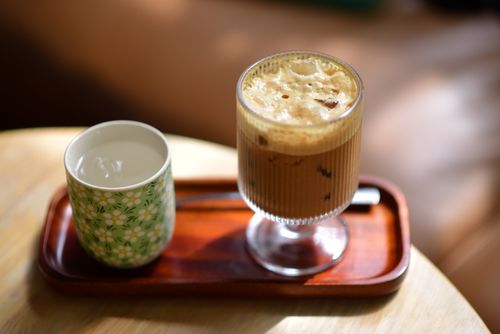 Free A refreshing iced coffee with frothy top beside a floral cup of water on a wooden tray in sunlight. Stock Photo