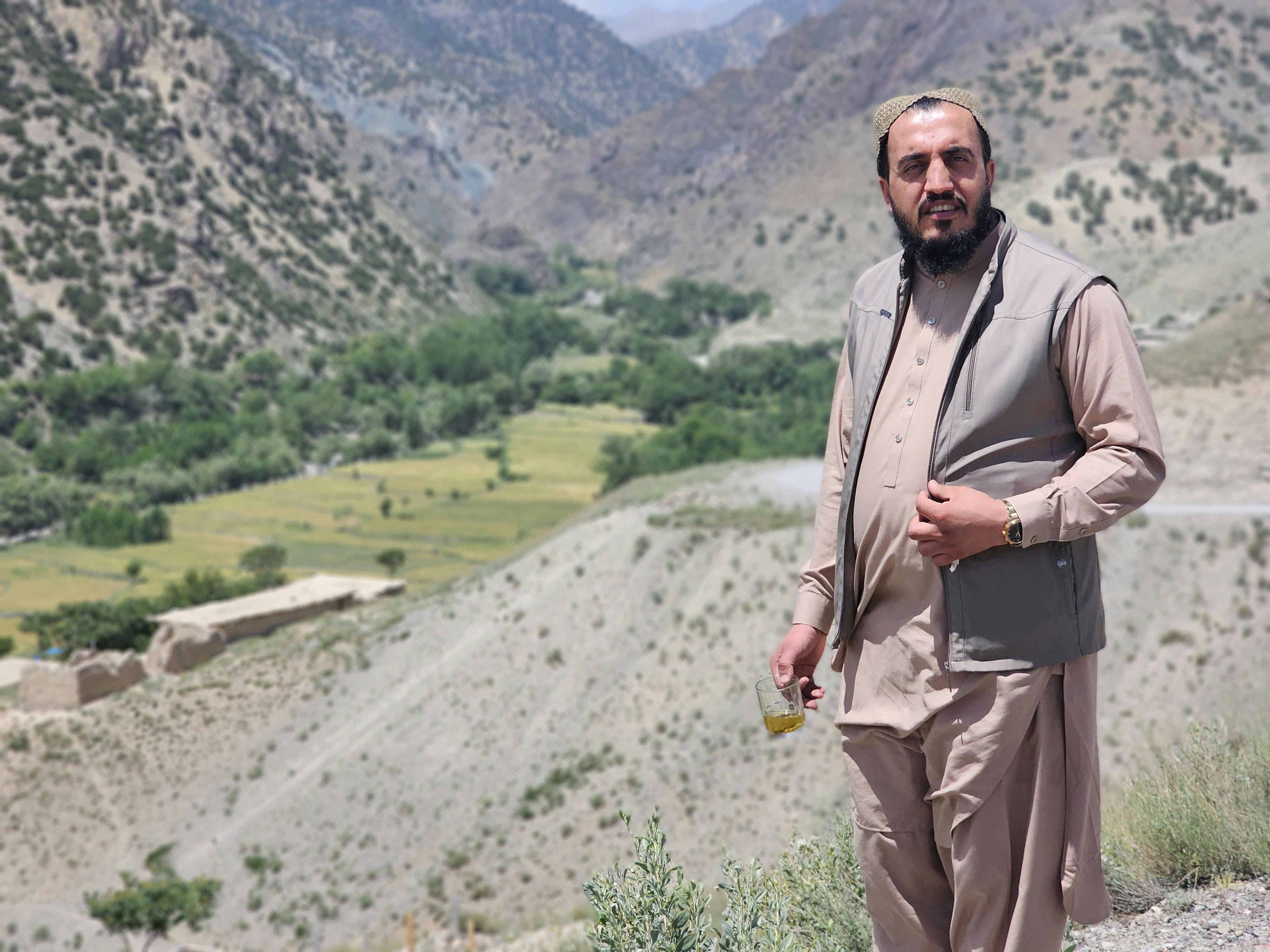 A man in traditional attire stands by a scenic mountainous landscape, holding a drink.