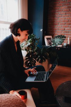 Focused woman works on a laptop while holding a coffee cup in a cozy home interior.