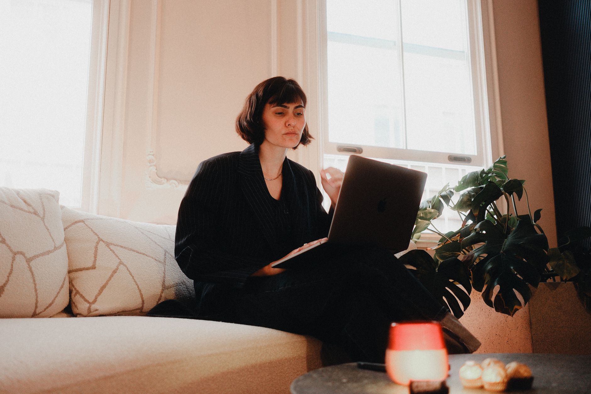 Woman in suit using laptop on sofa in bright room, modern lifestyle.