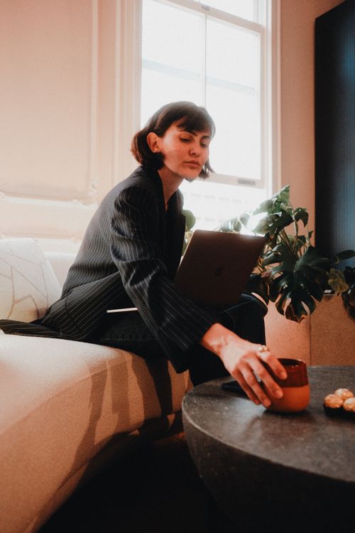 Free Focused woman in a stylish suit works on a laptop, reaching for coffee. Stock Photo