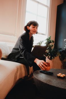Young woman in a cozy setting using laptop and enjoying a drink, exuding modern work-from-home vibes.