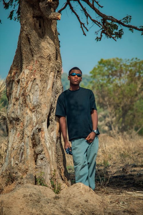 Free Casual young man wearing sunglasses leaning against a tree in a natural setting. Stock Photo