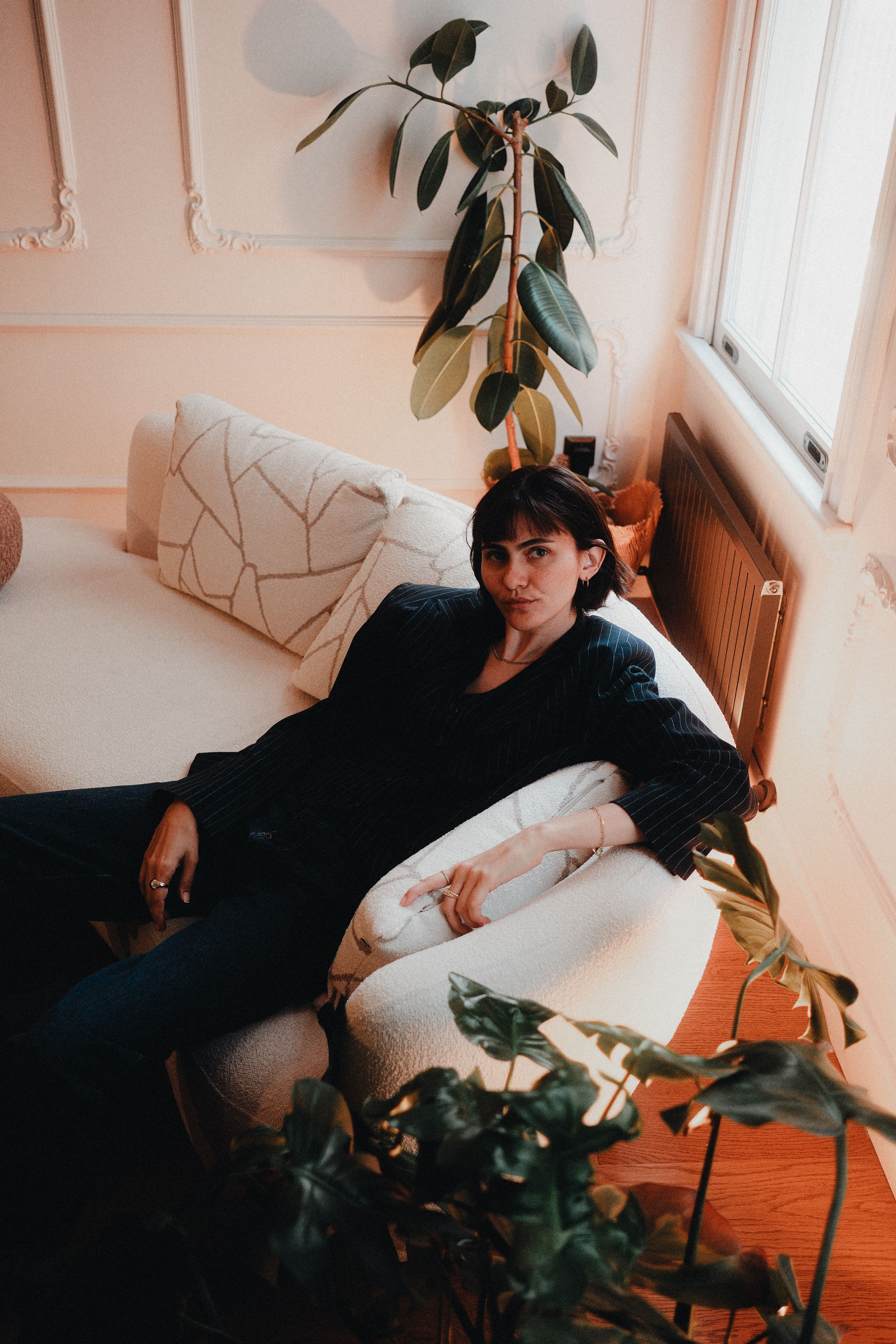 Woman relaxing on a sofa in a cozy, plant-filled living room with warm lighting.