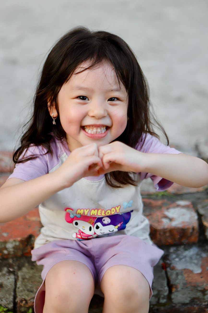 Smiling Vietnamese girl sitting outdoors making a heart shape with hands.