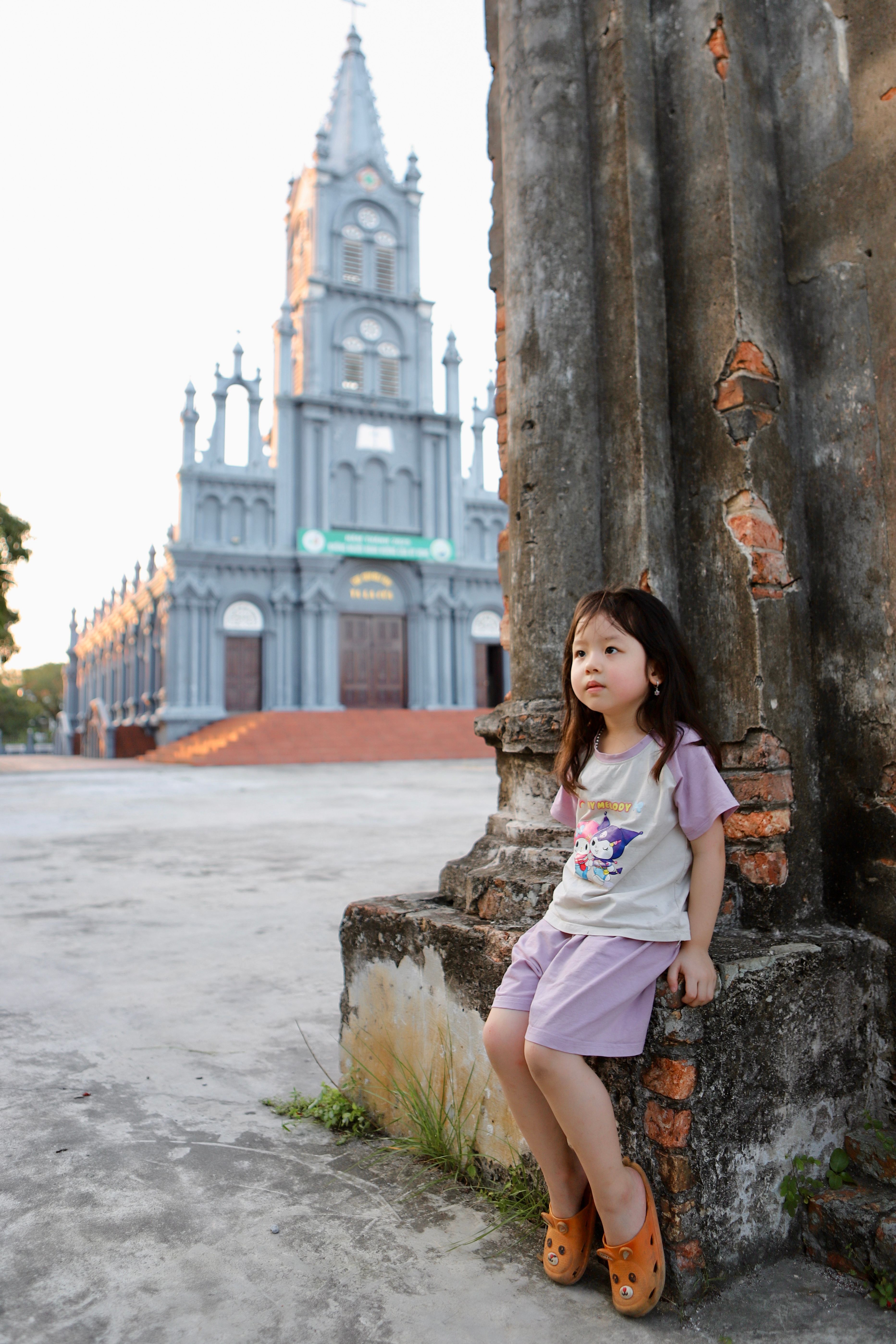 Young girl in purple outfit seated near a historic church in Hai Duong, Vietnam at dusk.