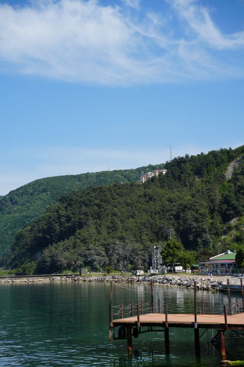 Free Tranquil lakeside scene featuring a wooden pier, calm waters, and lush green hills under a clear blue sky. Stock Photo