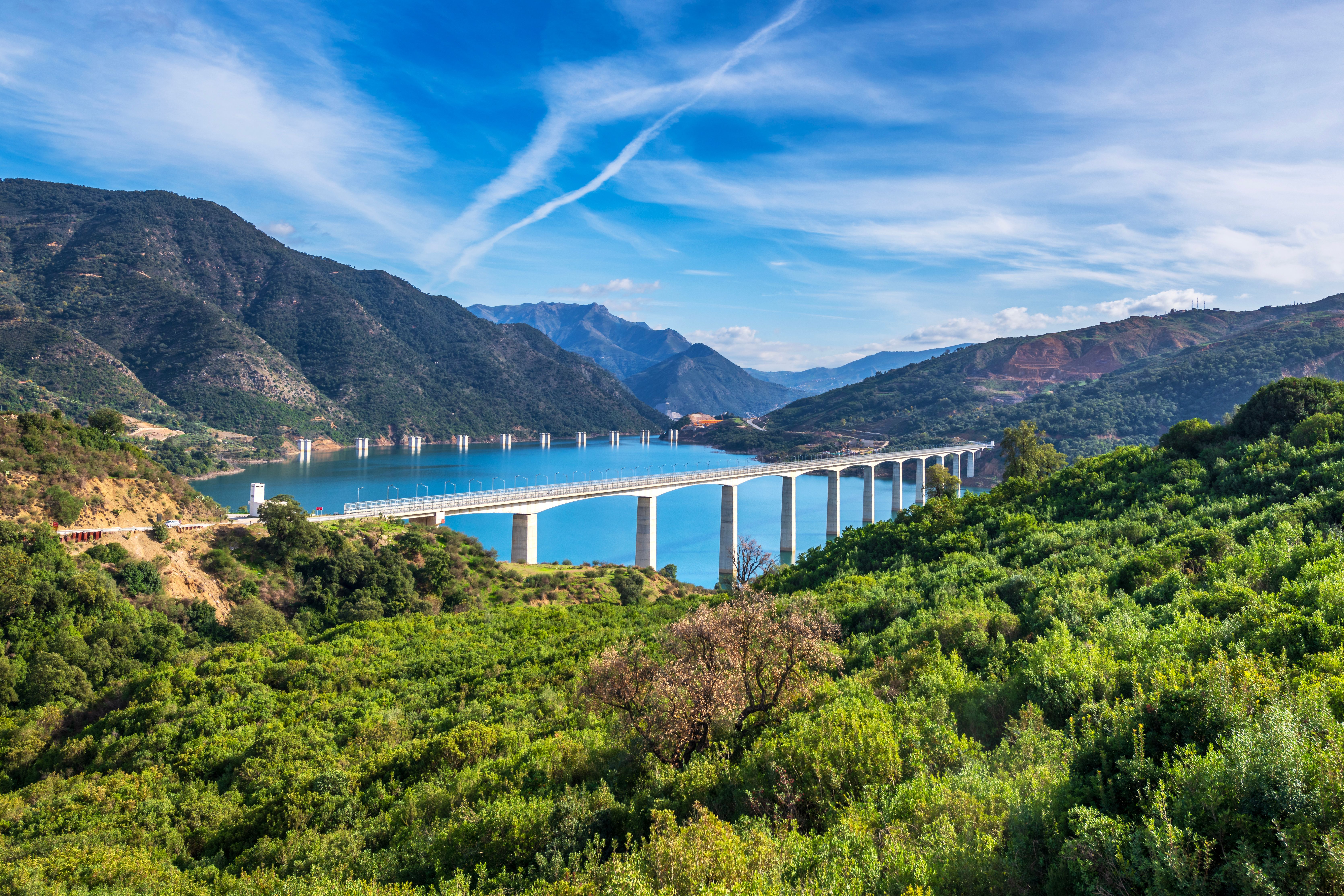A beautiful bridge spans a serene blue lake surrounded by lush greenery and mountain ranges under a clear sky.