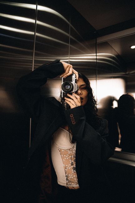 Stylish woman taking a photo with a vintage camera in an elevator with reflective walls.