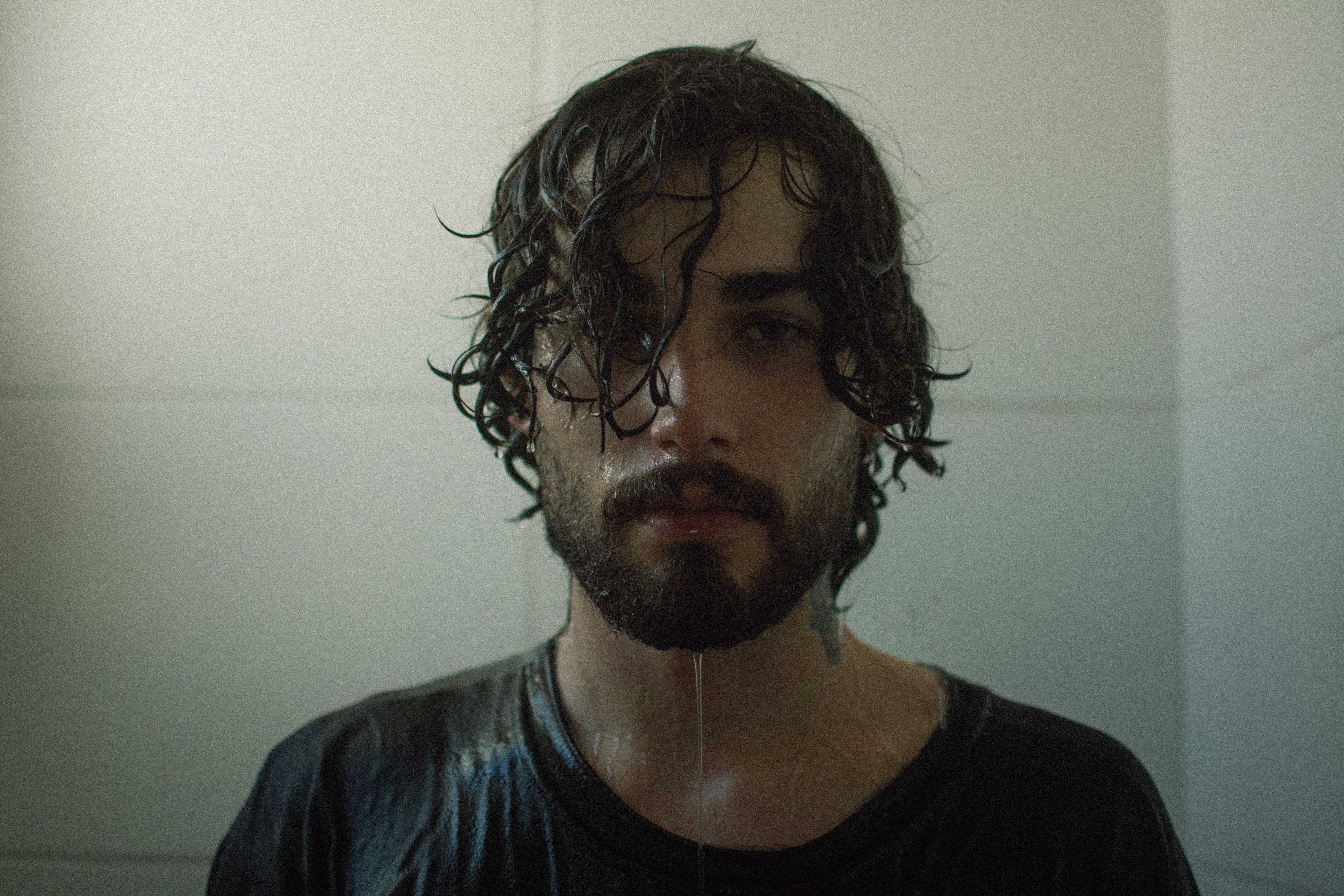 A moody, introspective portrait of a man with wet hair standing in a shower.