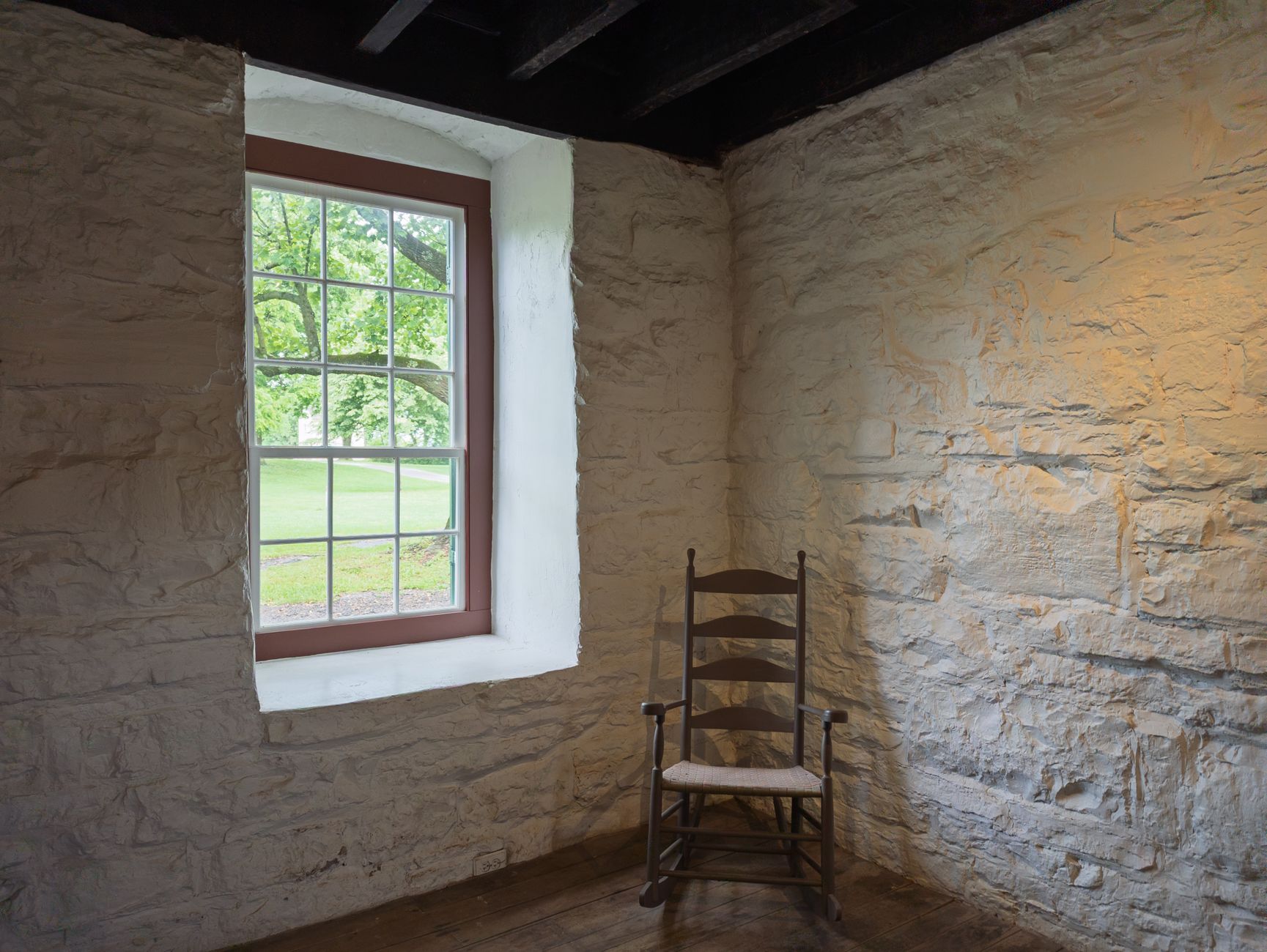 A peaceful scene of an antique chair beside a window in a Shaker-style stone room.