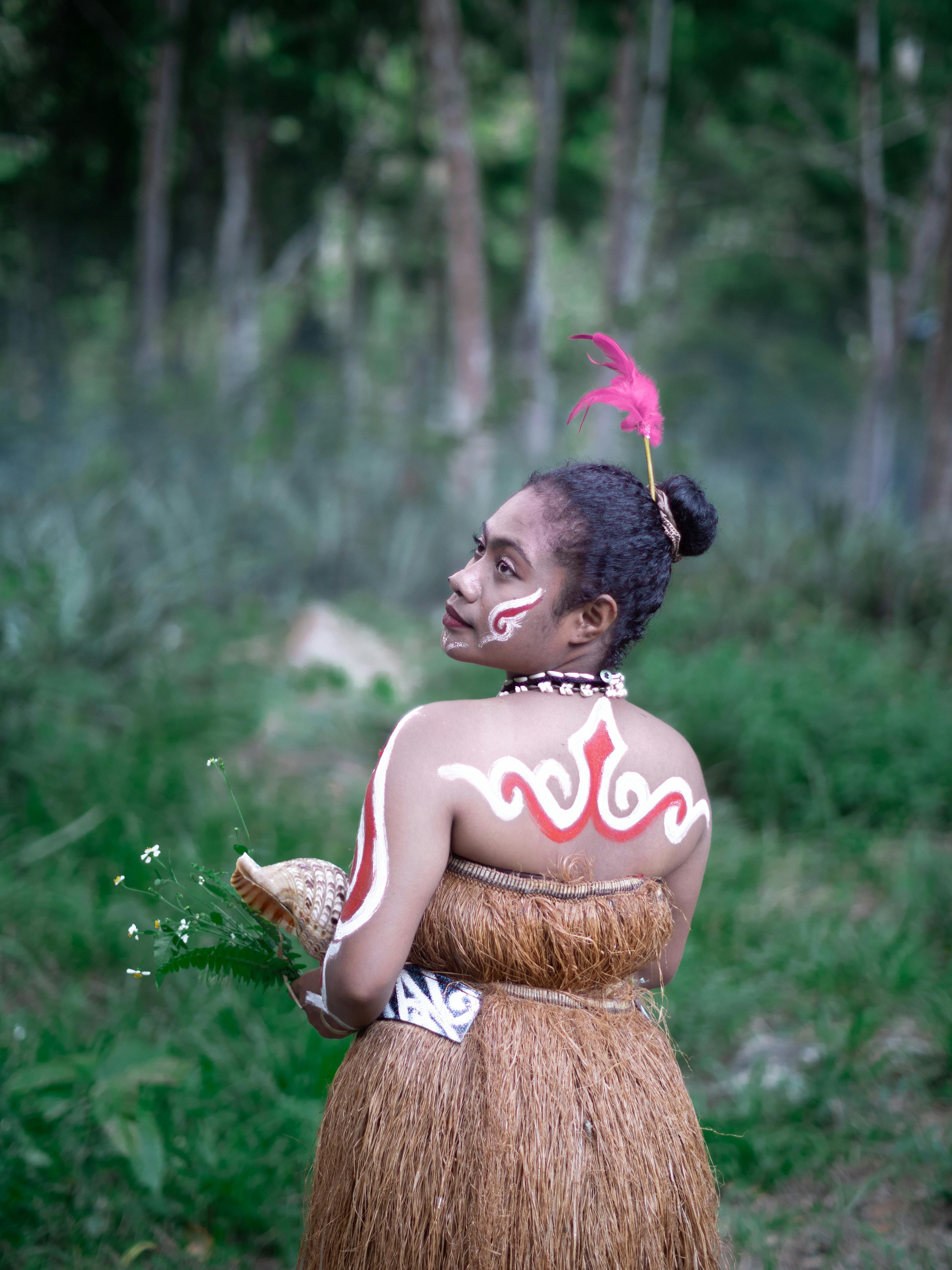 A woman in traditional tribal attire with body paint stands in a lush forest setting.
