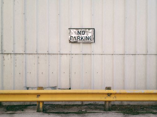 Free A rustic metal 'No Parking' sign on an industrial wall with a yellow barrier. Stock Photo