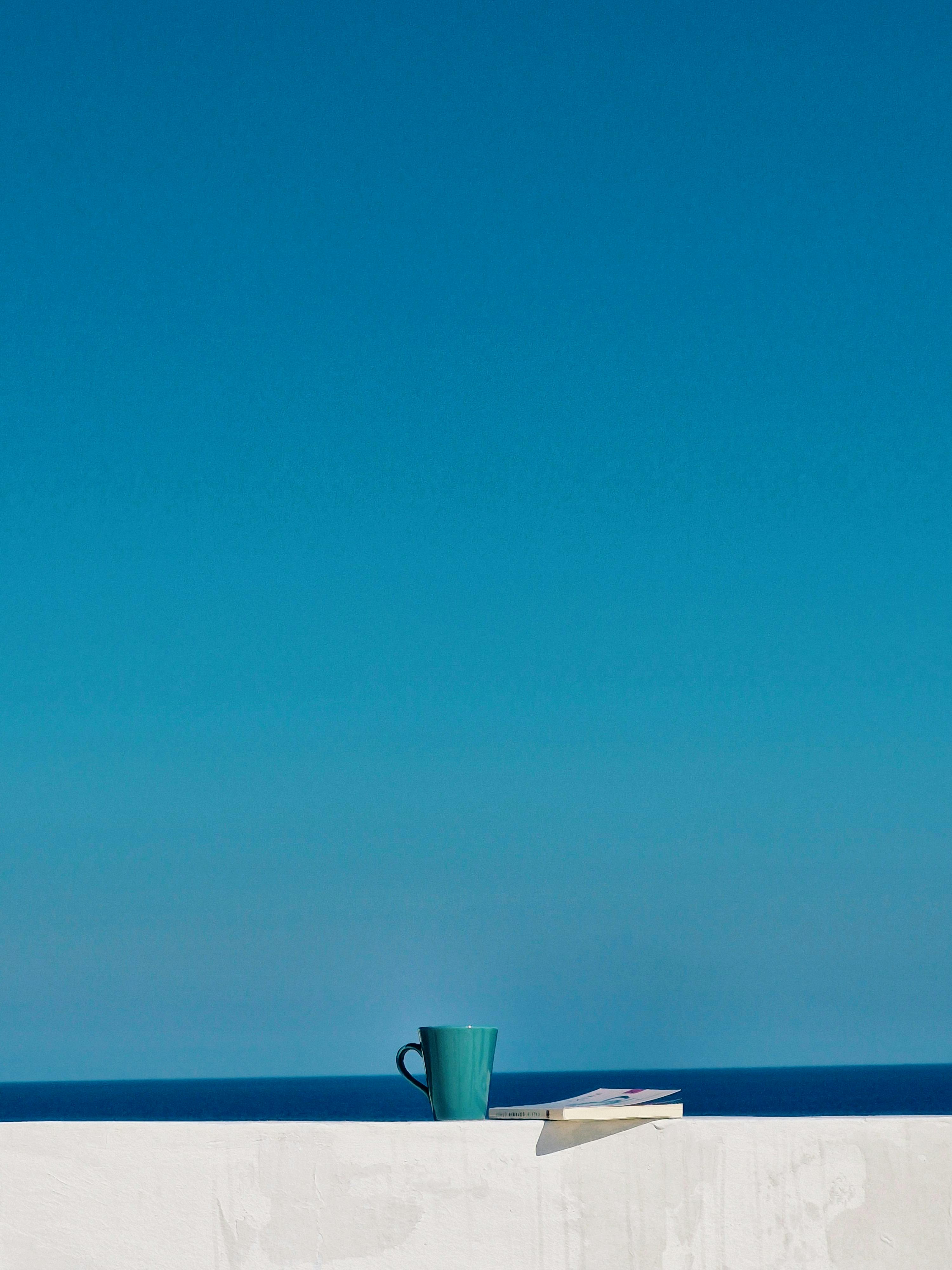 Enjoy a serene moment with a coffee and book by the azure sea under a clear blue sky.