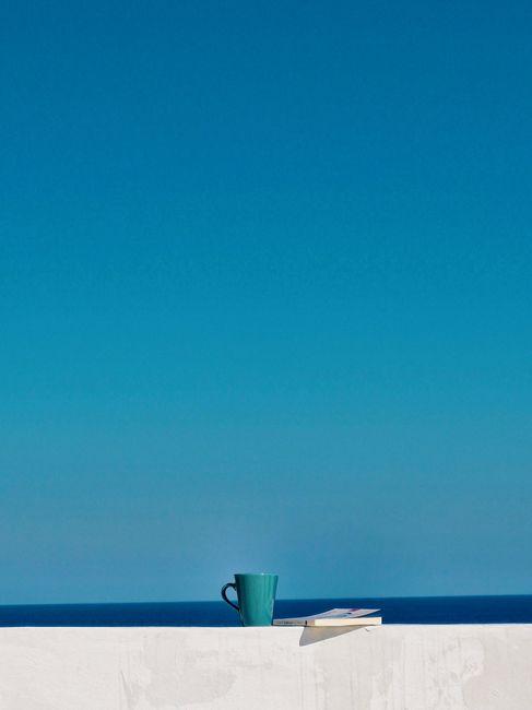Enjoy a serene moment with a coffee and book by the azure sea under a clear blue sky.