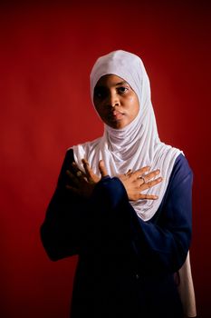 A woman in a white hijab and blue clothing poses against a red backdrop.