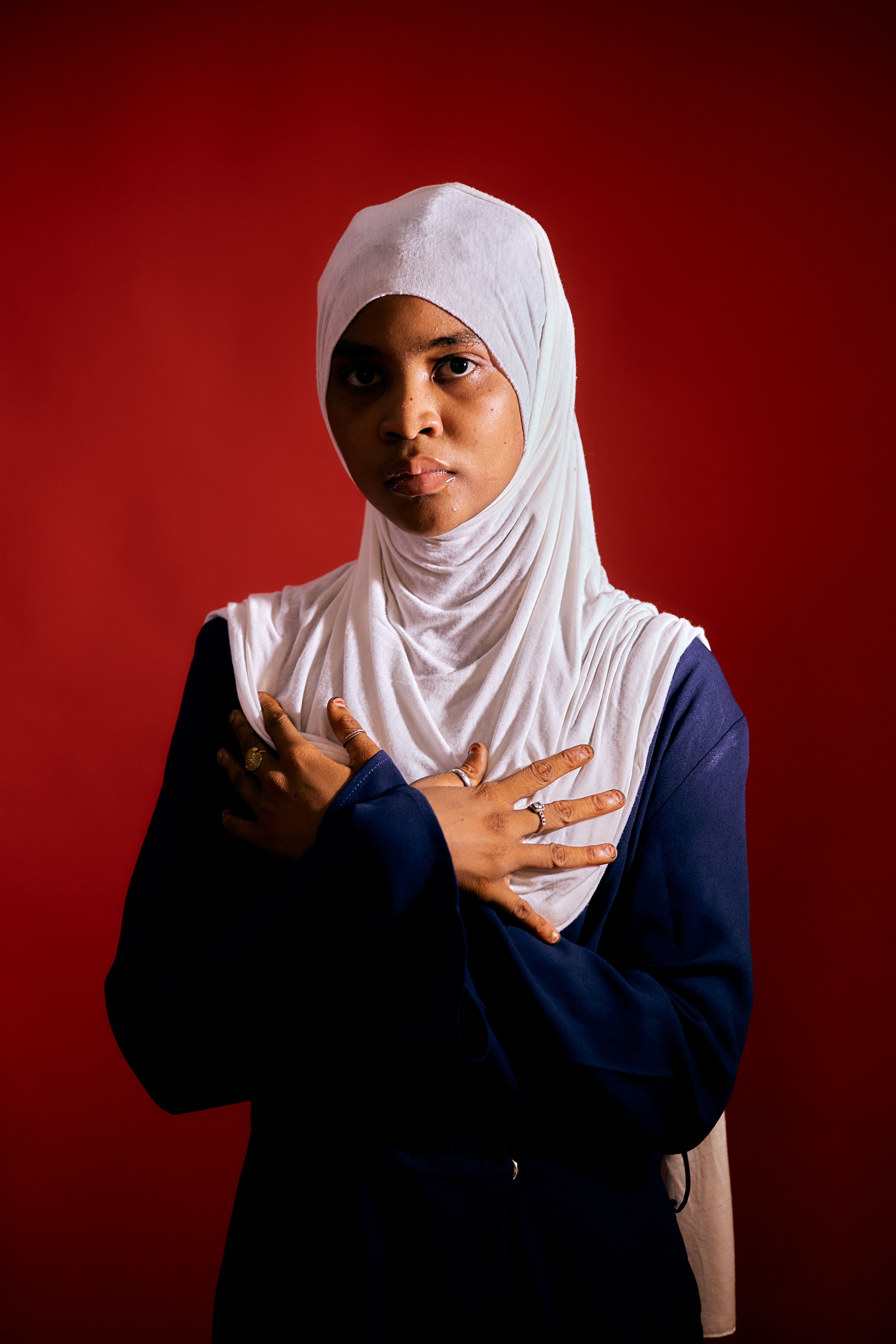 A thoughtful woman in a white hijab poses confidently against a vibrant red backdrop.