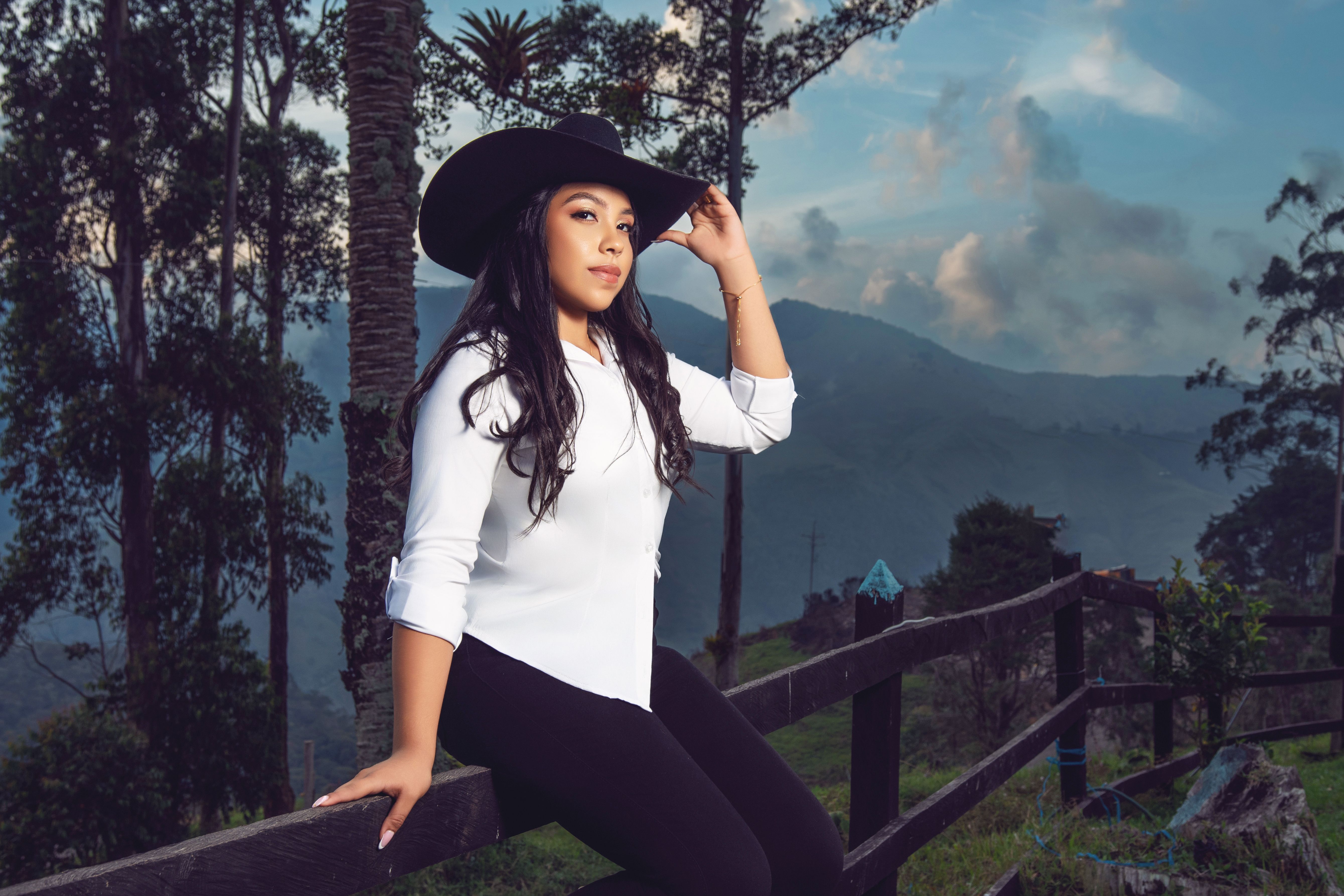 Woman in a black hat and white shirt posing in a scenic outdoor setting with mountains and trees.