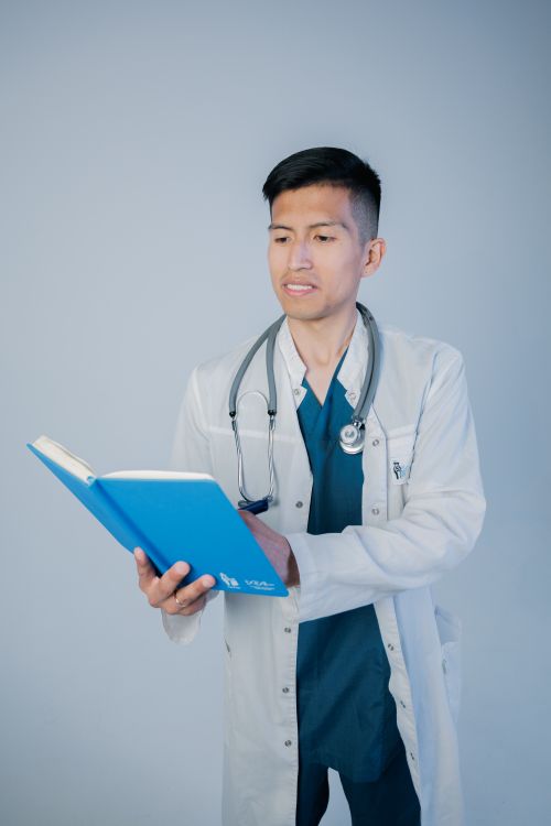 Free A young male doctor in a white coat reads a blue medical book against a plain studio background. Stock Photo