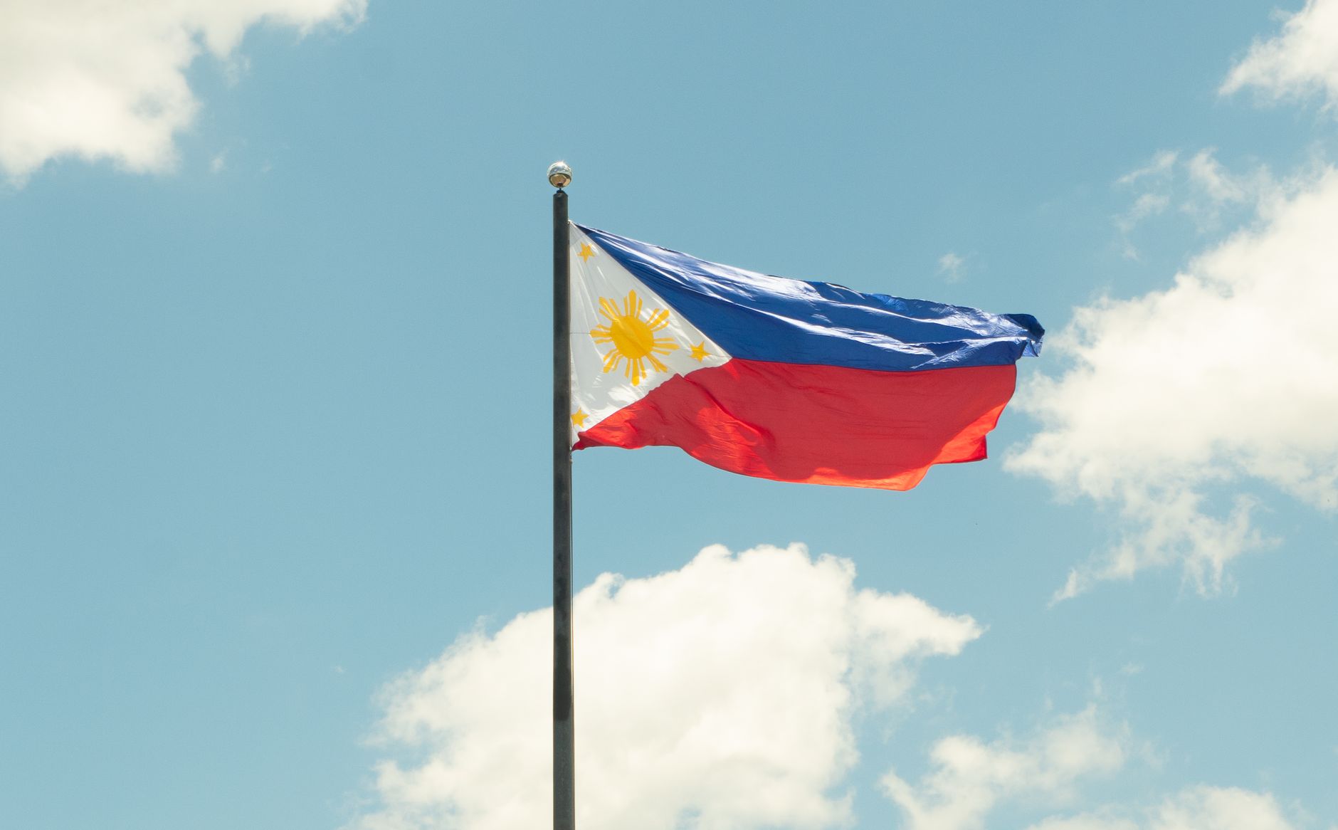 The national flag of the Philippines waving proudly in a clear blue sky with clouds.