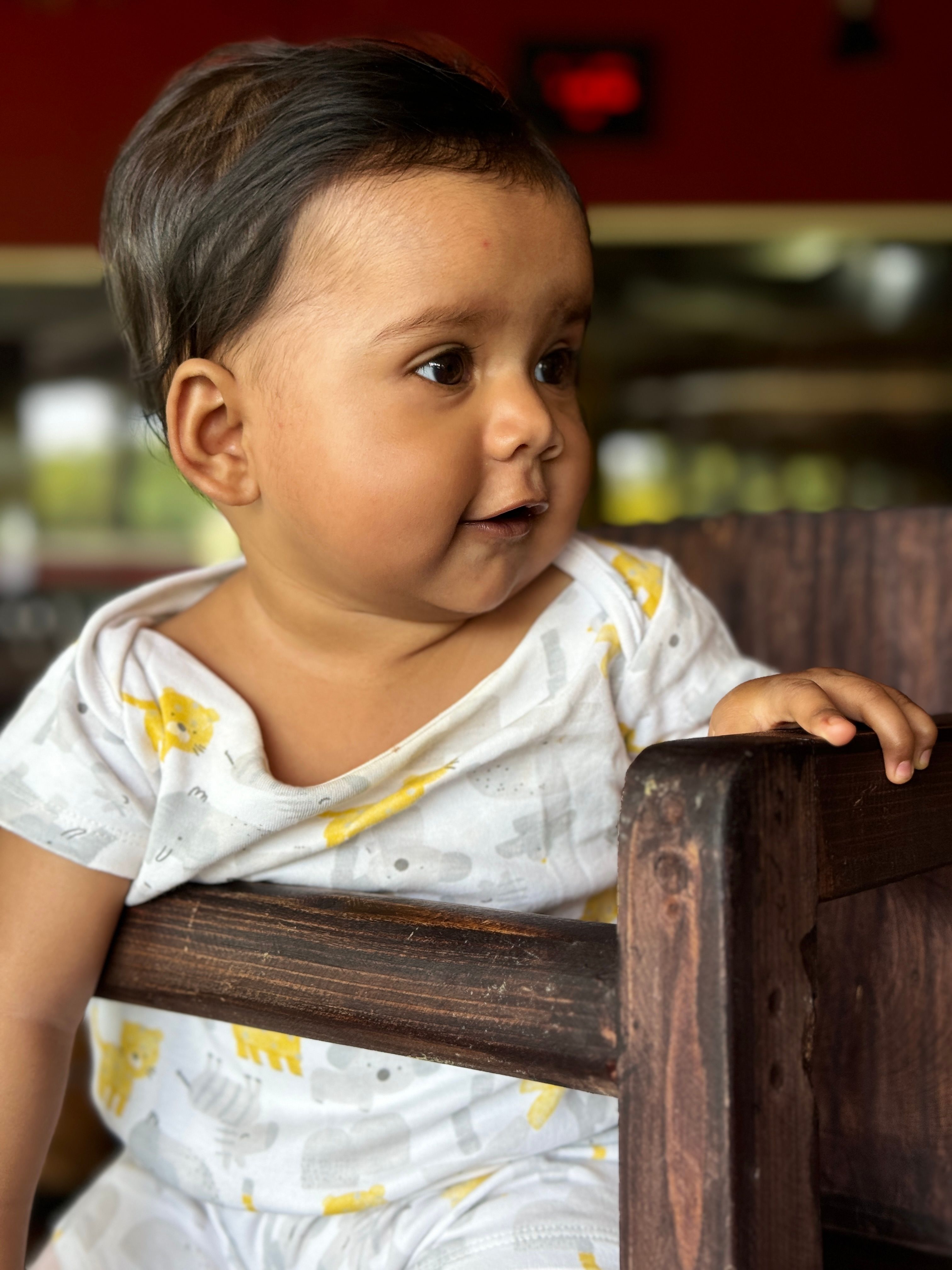 Charming baby in printed outfit sitting on a rustic wooden chair inside a cozy setting.