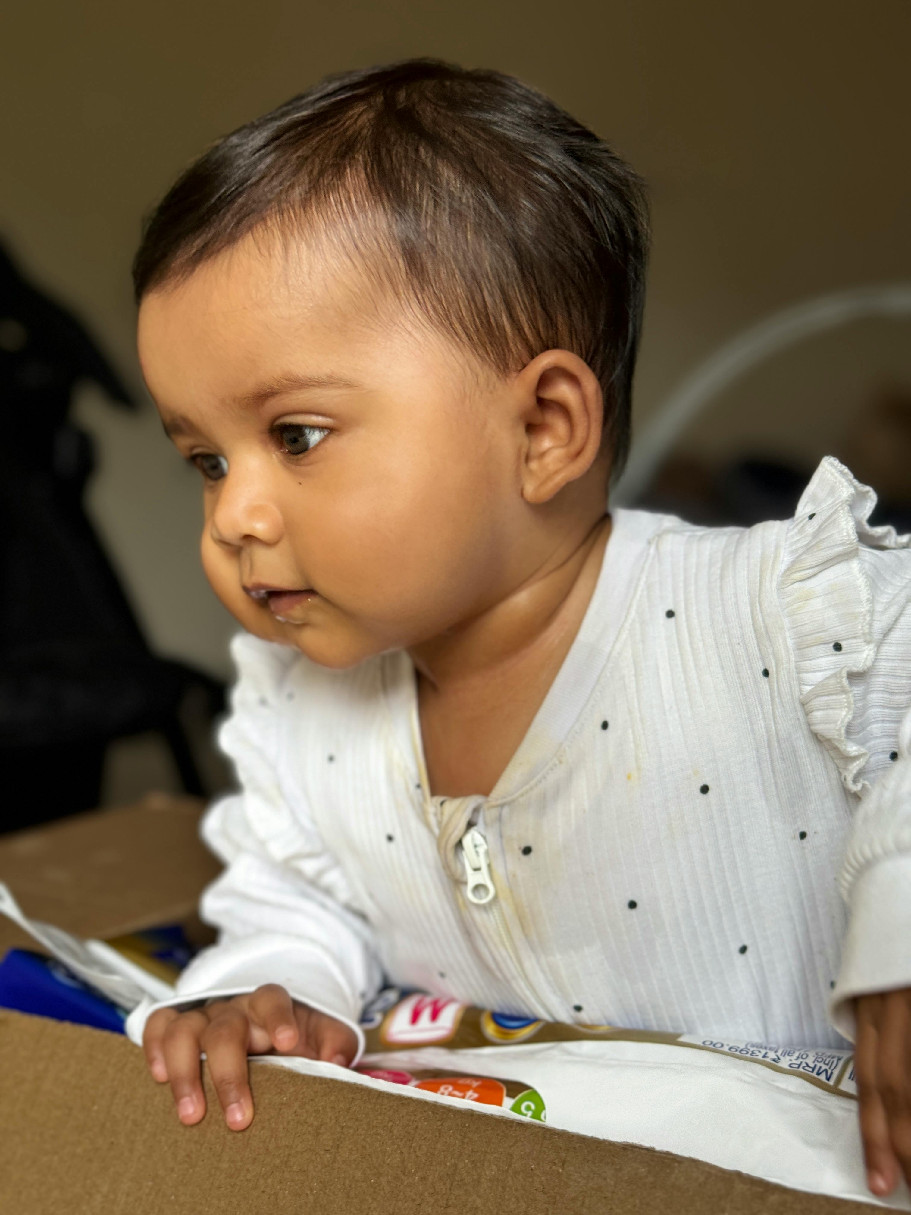 Cute baby in a white onesie exploring a cardboard box indoors, captured in a candid moment.