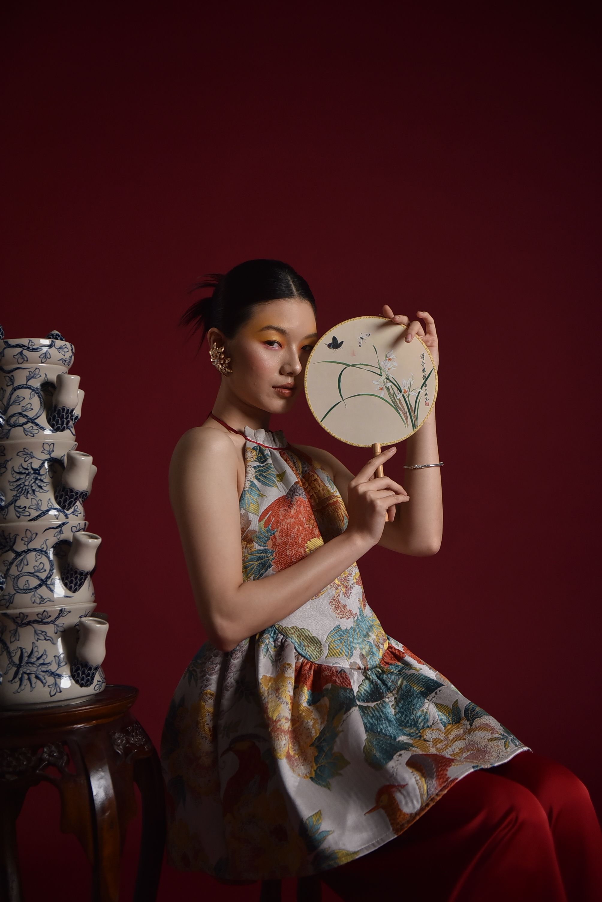 Stylish woman in floral dress holding a decorative fan in an artistic studio setting.
