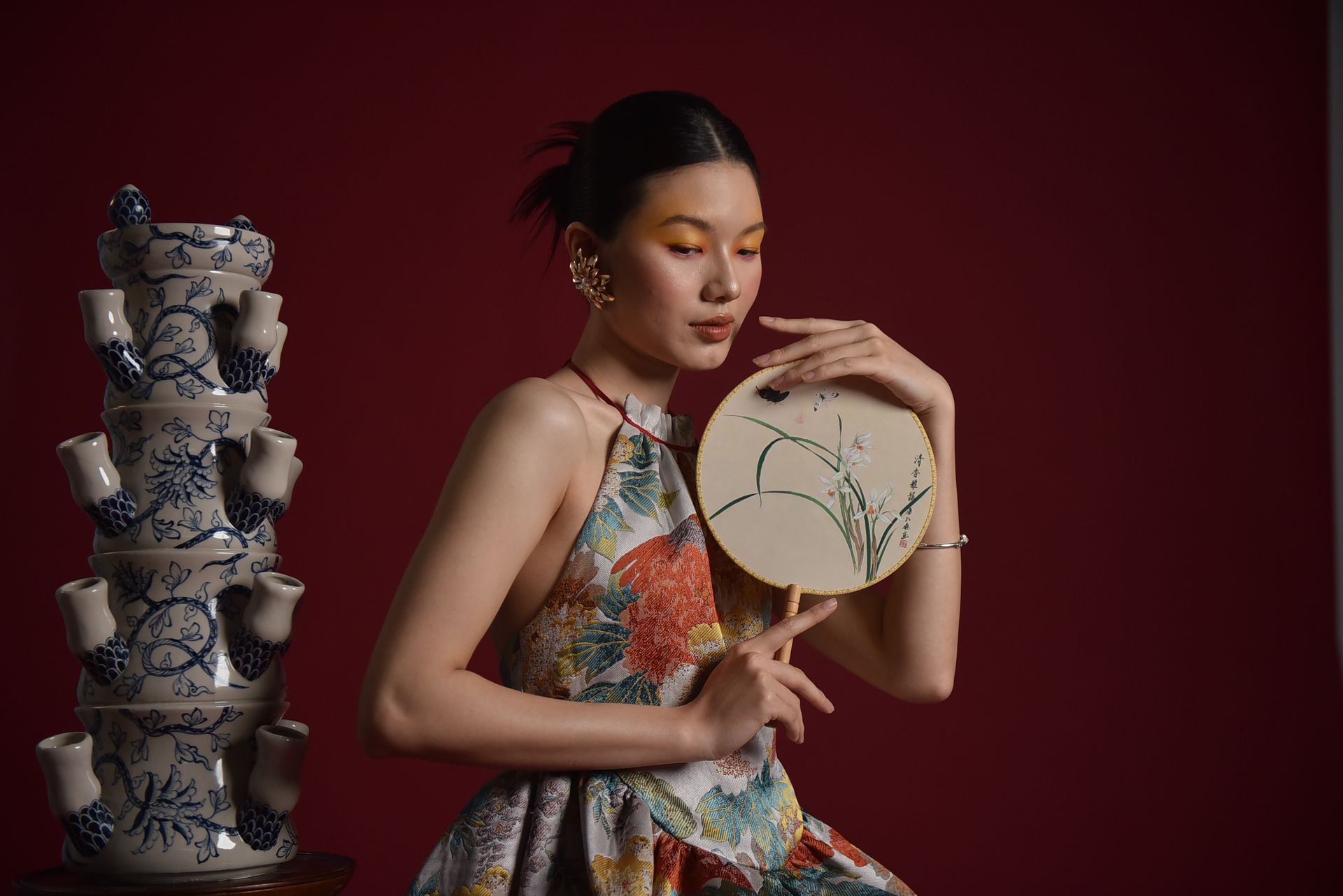 Portrait of a stylish woman in floral dress holding ornate fan beside ceramic art.