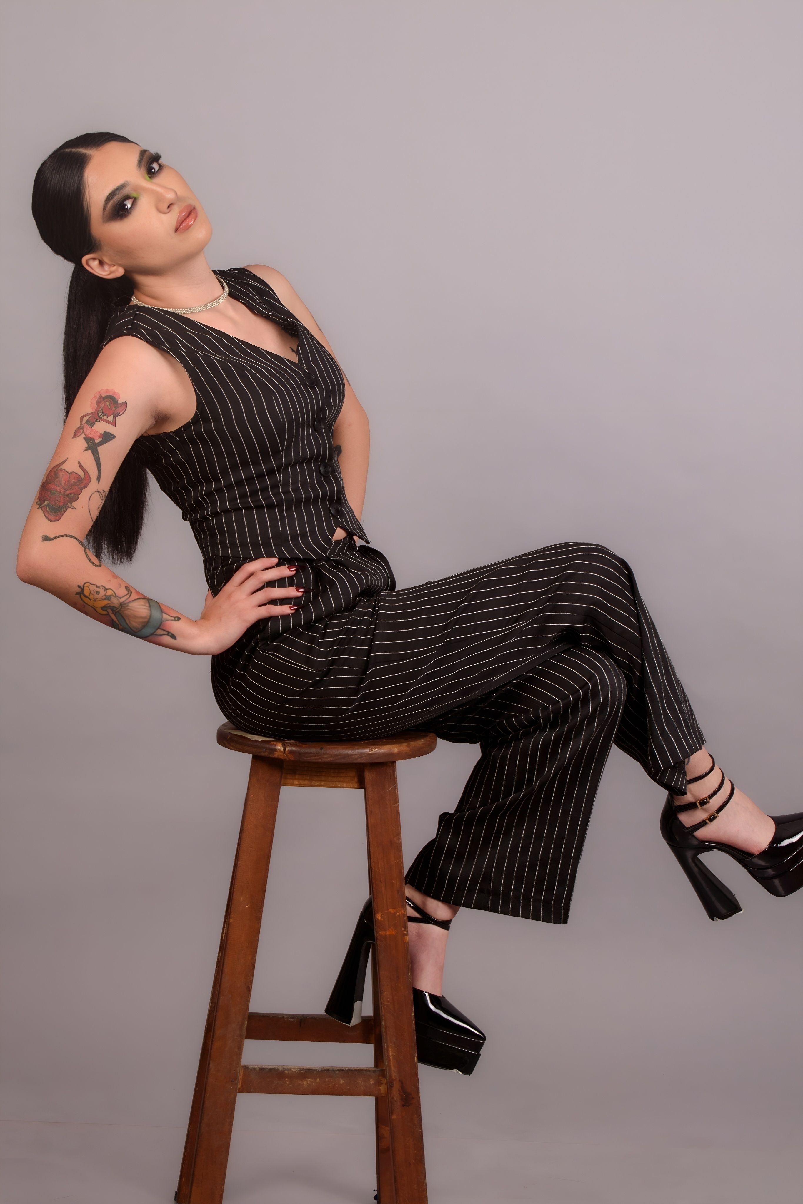 Stylish woman in pinstripe suit with tattoos sitting on stool, showcasing elegance and attitude.