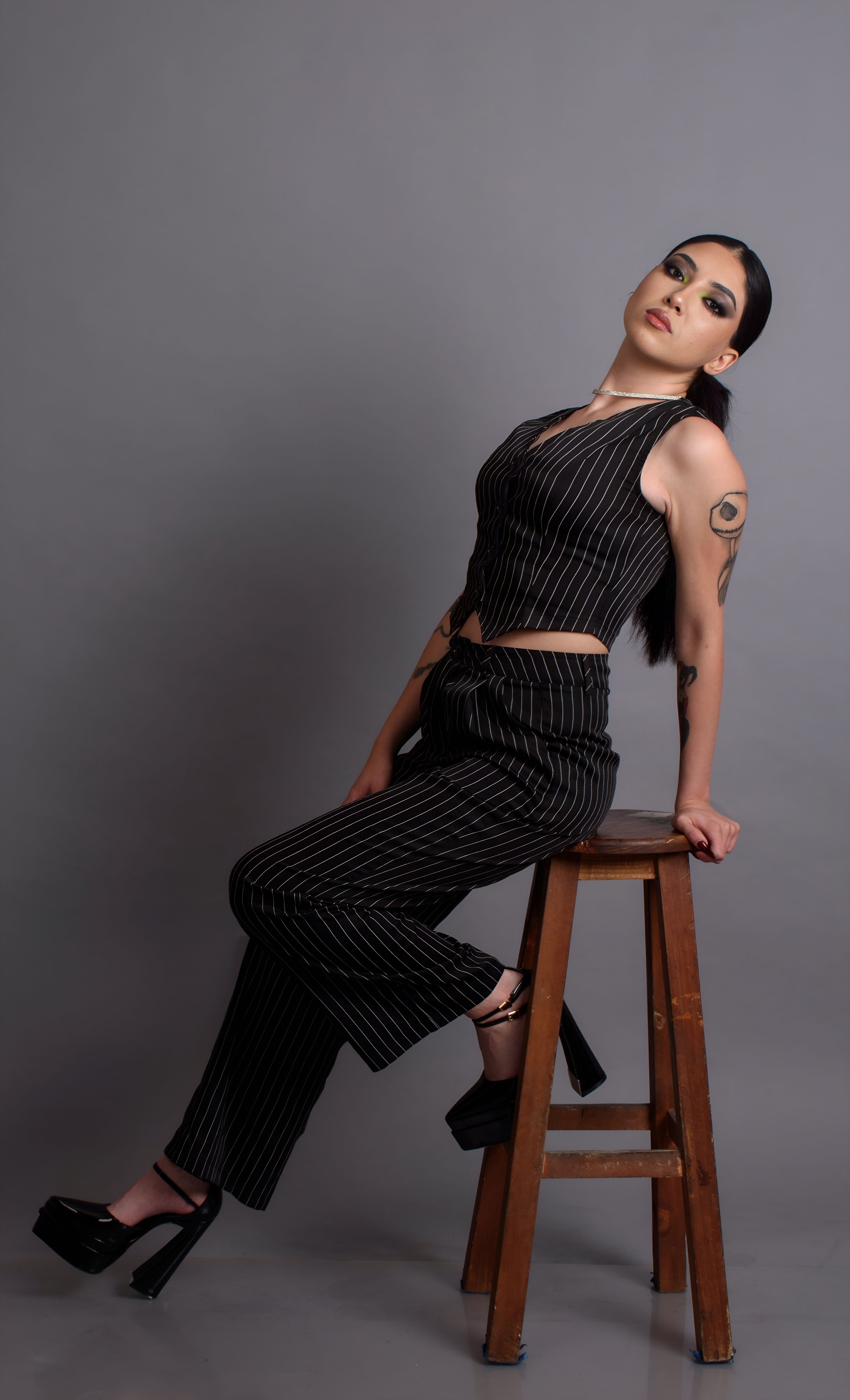 Mulher elegante em um traje listrado posa com confiança em um banquinho, exibindo moda moderna e tatuagens únicas.