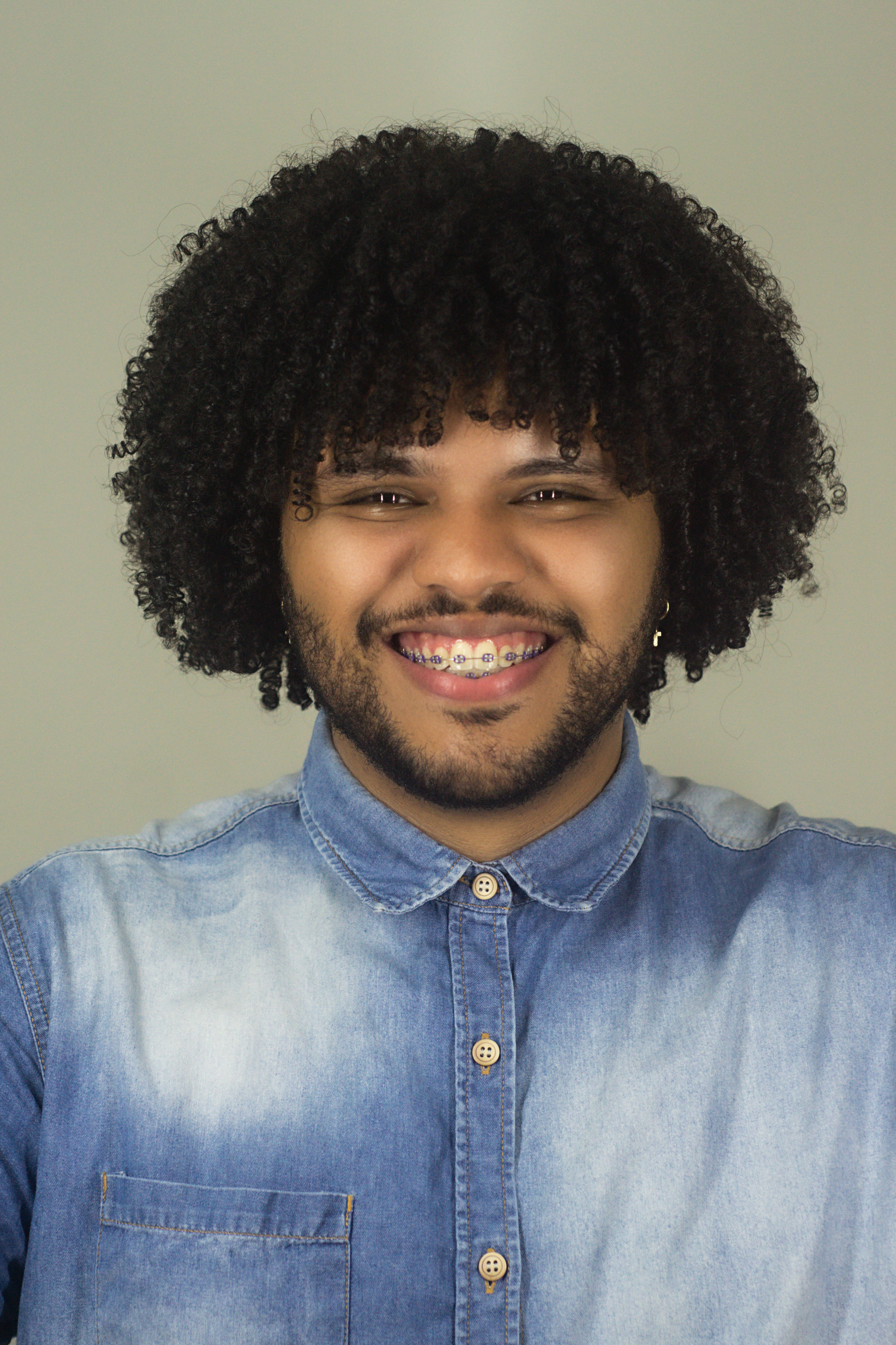 Portrait of a young man with curly hair and braces, smiling warmly in a denim shirt.
