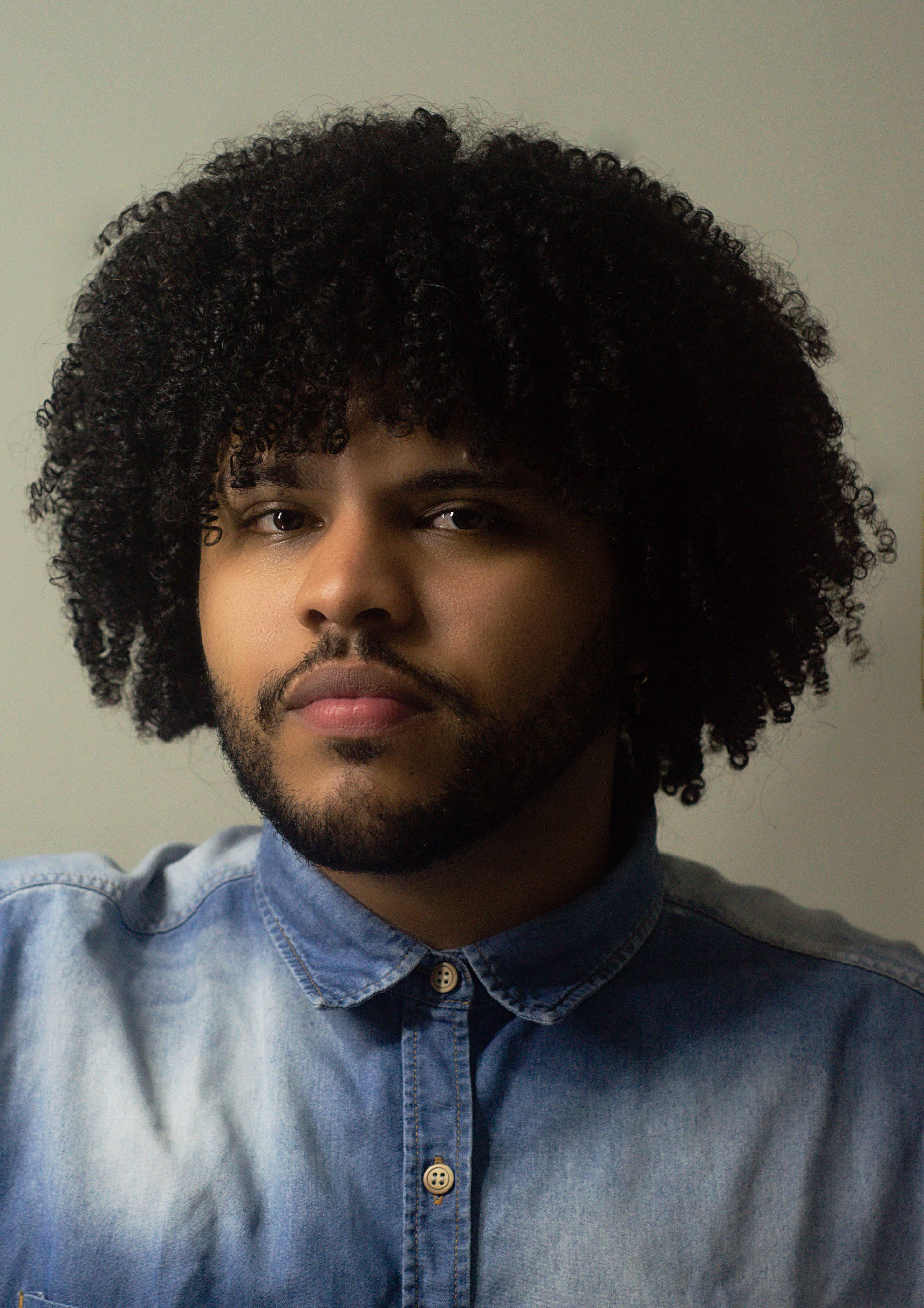 Close-up portrait of a young man with curly hair wearing a denim shirt.