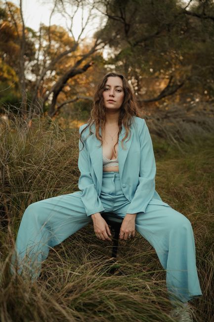 Confident young woman in a blue suit sits surrounded by nature, exuding elegance.