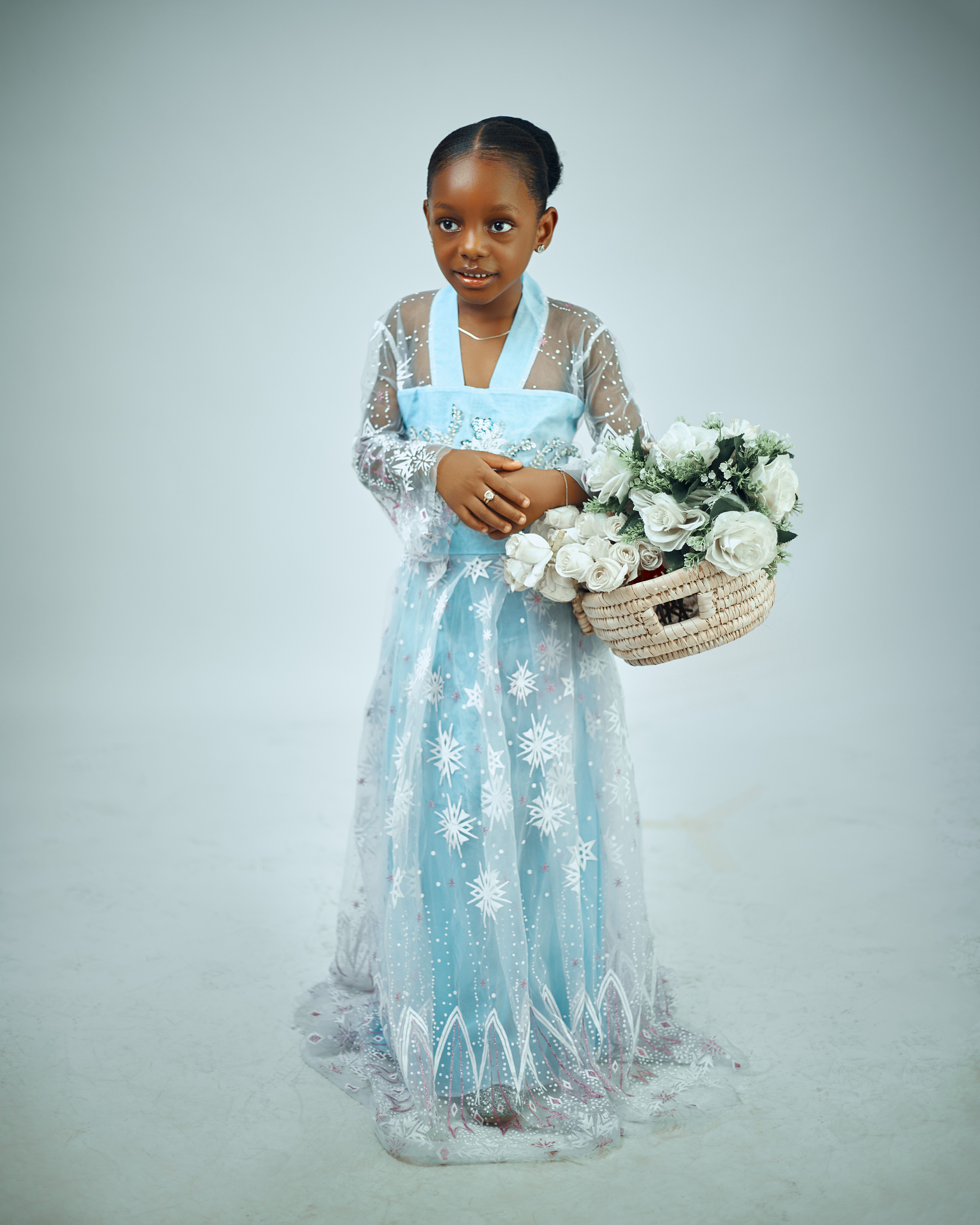 Young girl in elegant blue dress carrying a basket of white flowers against a neutral backdrop.