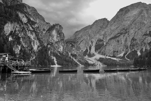 Peaceful black and white scene of rowboats on a lake surrounded by majestic mountains.