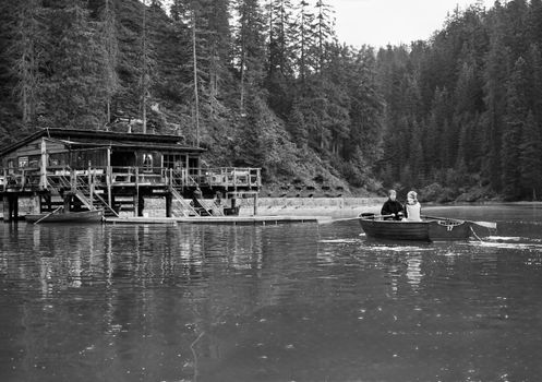 Black and white photo of a couple boating by a rustic cabin in a forested area.