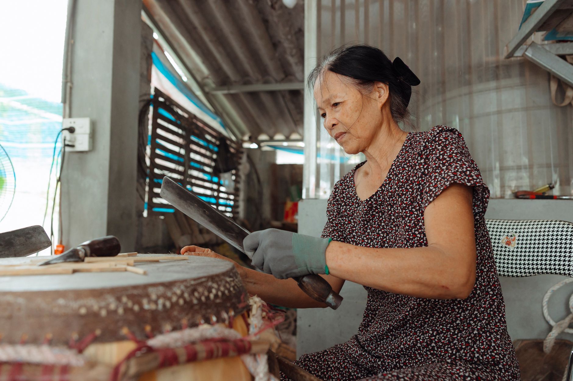 An elderly woman crafting a drum blade in a rustic indoor workshop, focusing on traditional methods.