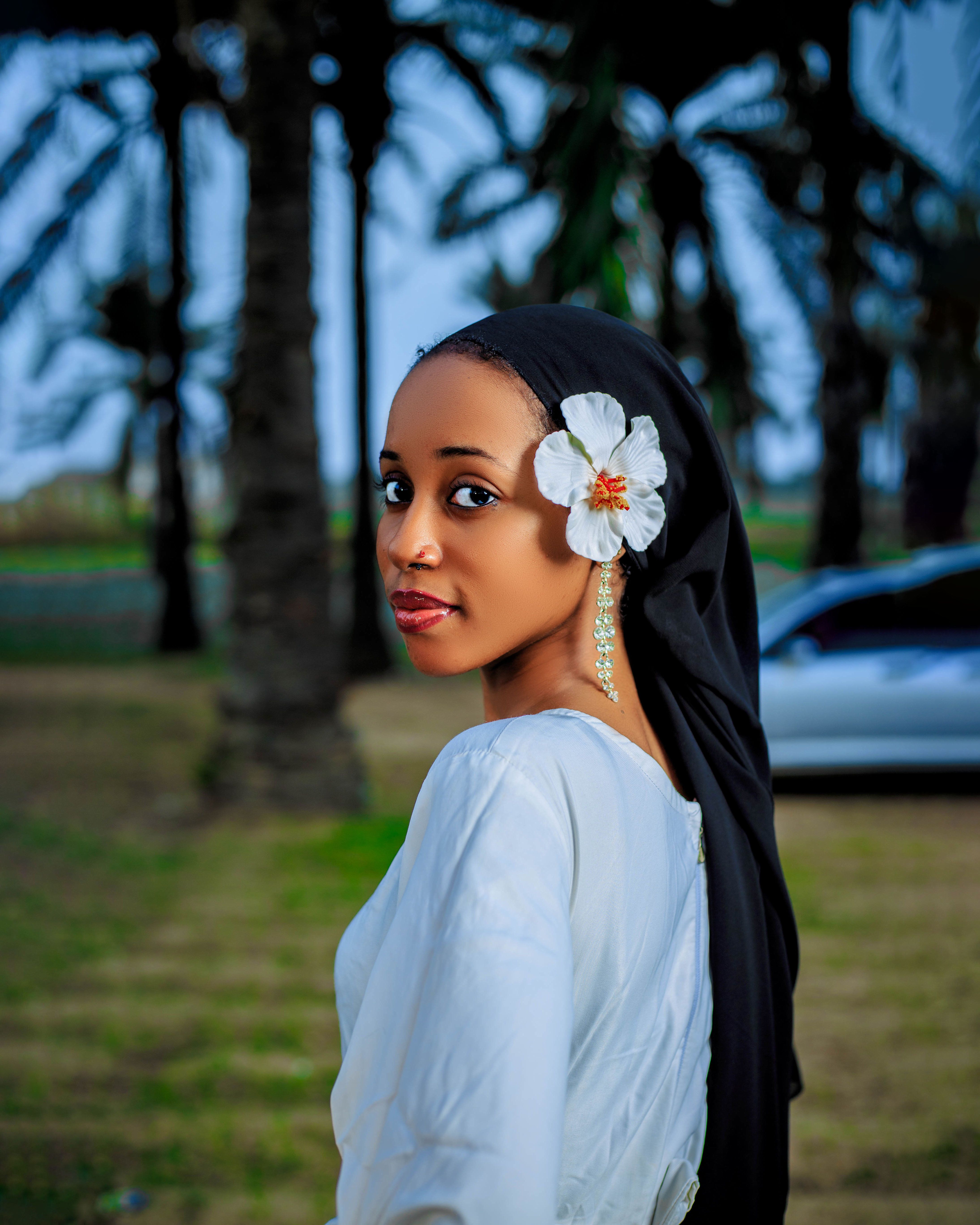 Graceful woman with white flower in hair, posing outdoors among palm trees, exuding elegance and charm.