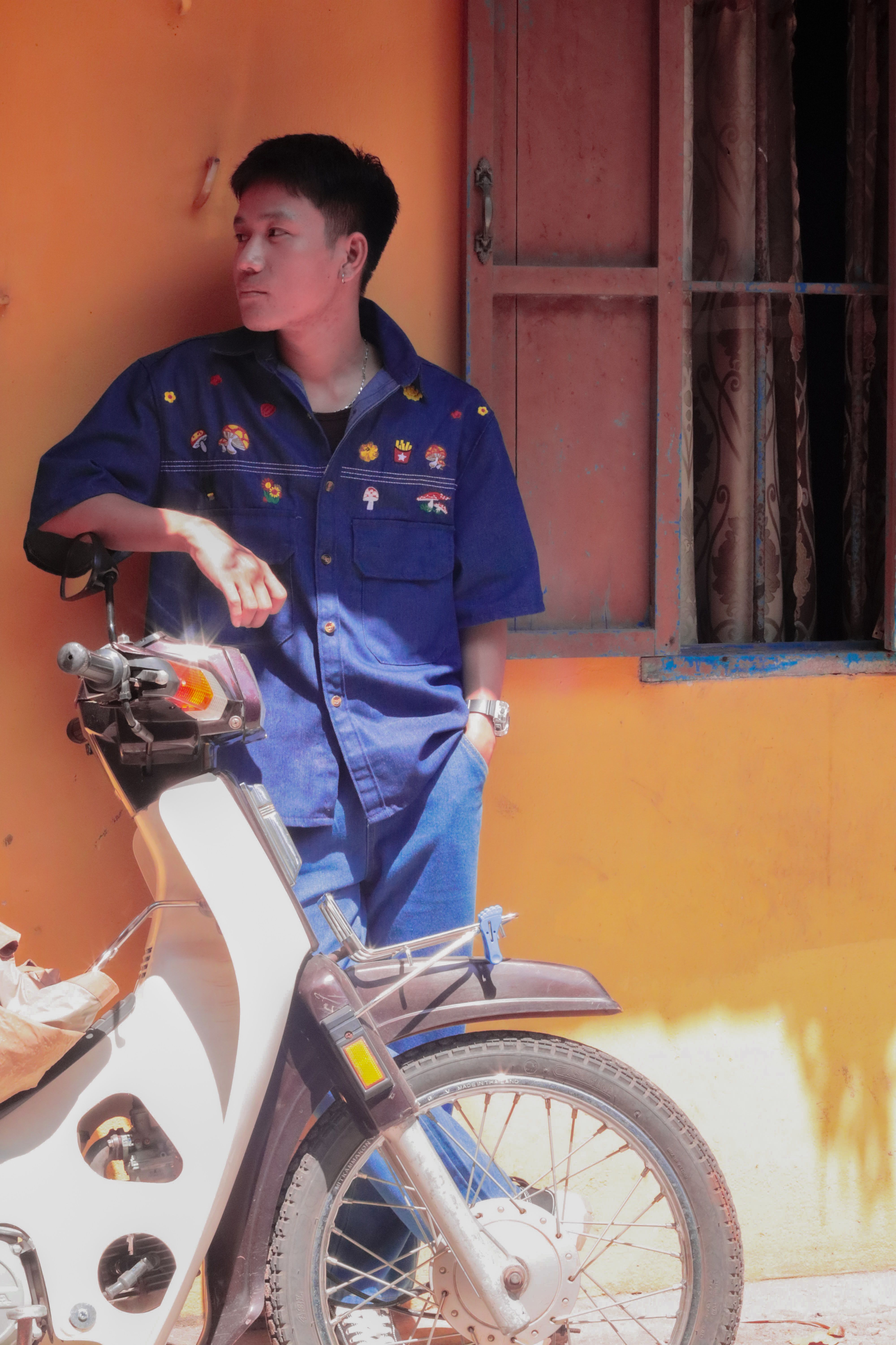 Young man in blue outfit leaning on motorcycle against a bright orange wall, reflecting urban lifestyle.