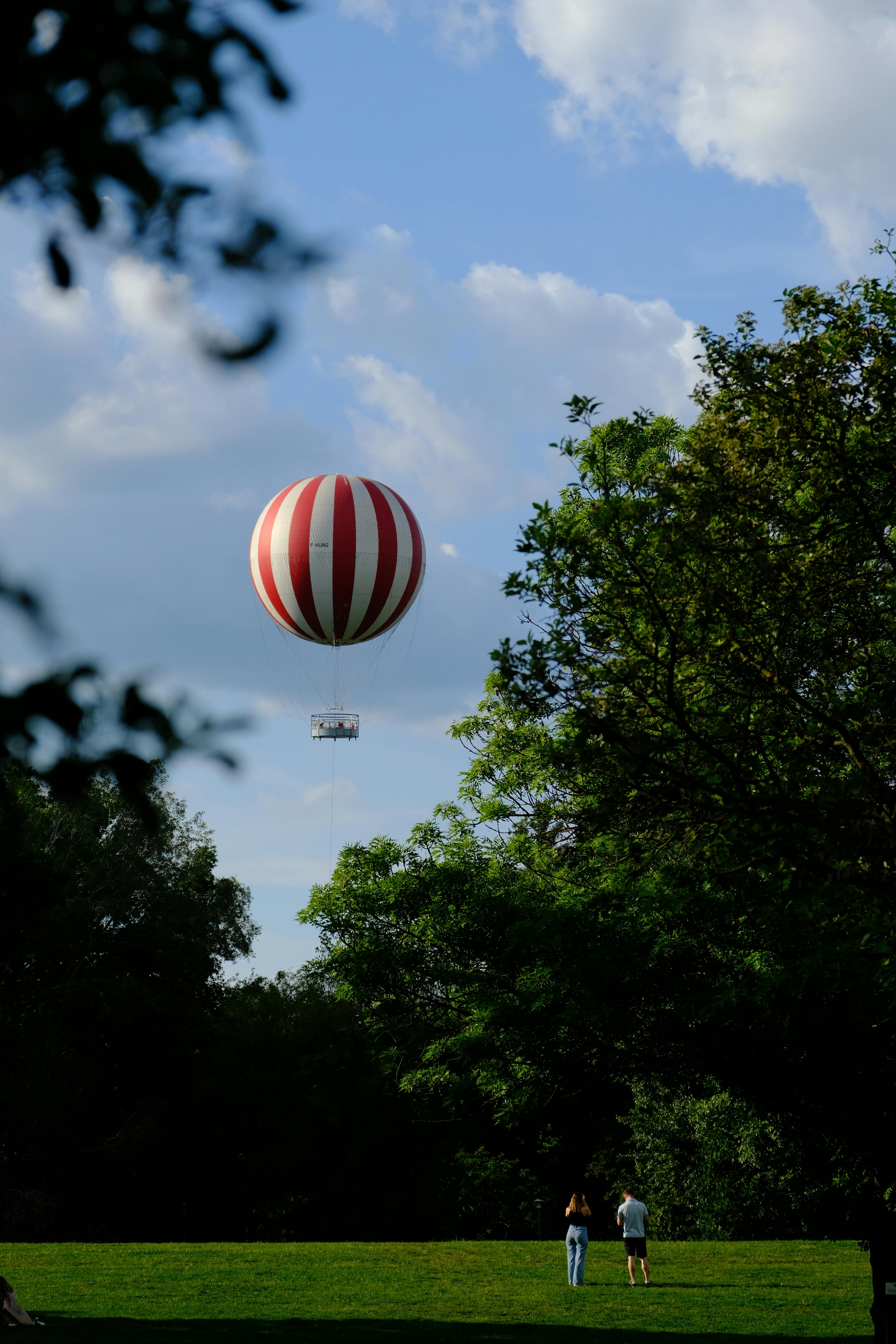 Red and white hot air balloon soaring above a vibrant green park with trees and people below.