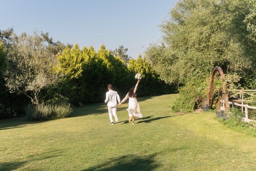 A couple in wedding attire joyfully running through a sunny garden setting.