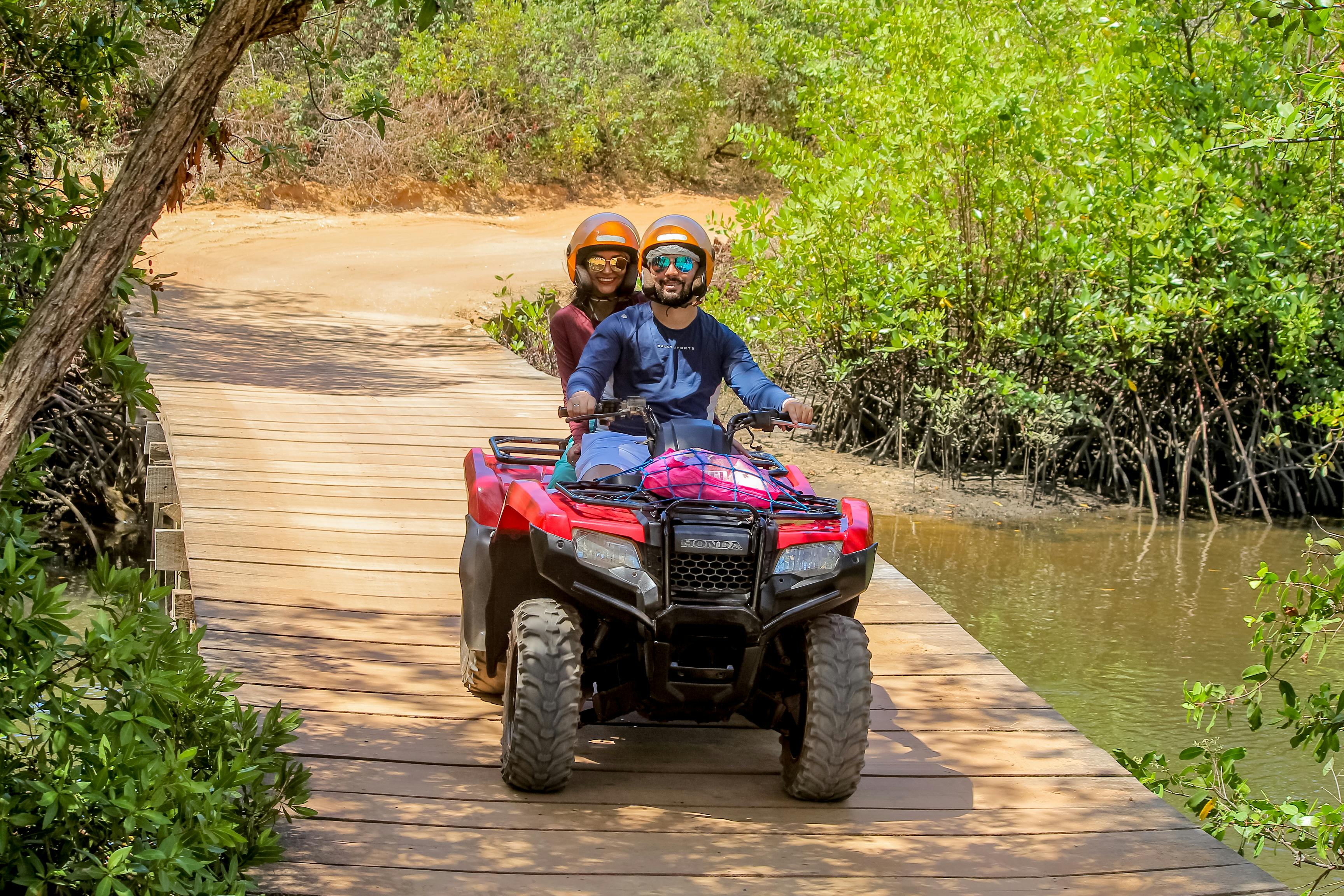 Couple riding an ATV on a wooden path surrounded by lush greenery during a sunny outdoor adventure.