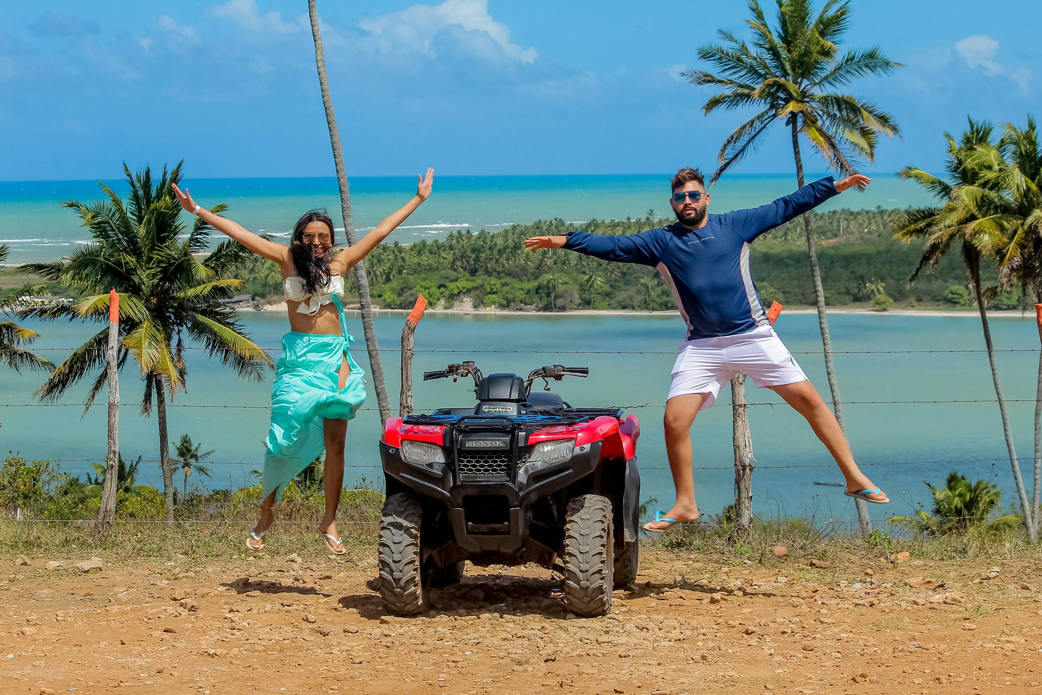 Two adults joyfully jumping beside an ATV on a tropical beach with clear skies and palm trees.